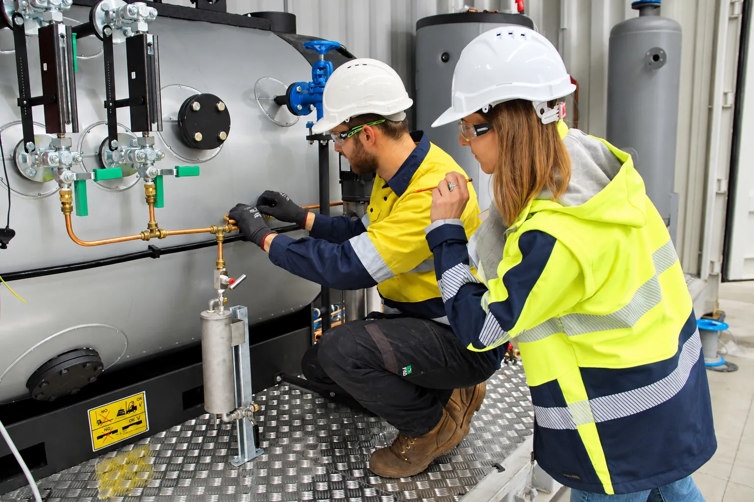 Boiler technician showing a trainee in hi-vis how to operate a valve on the boiler