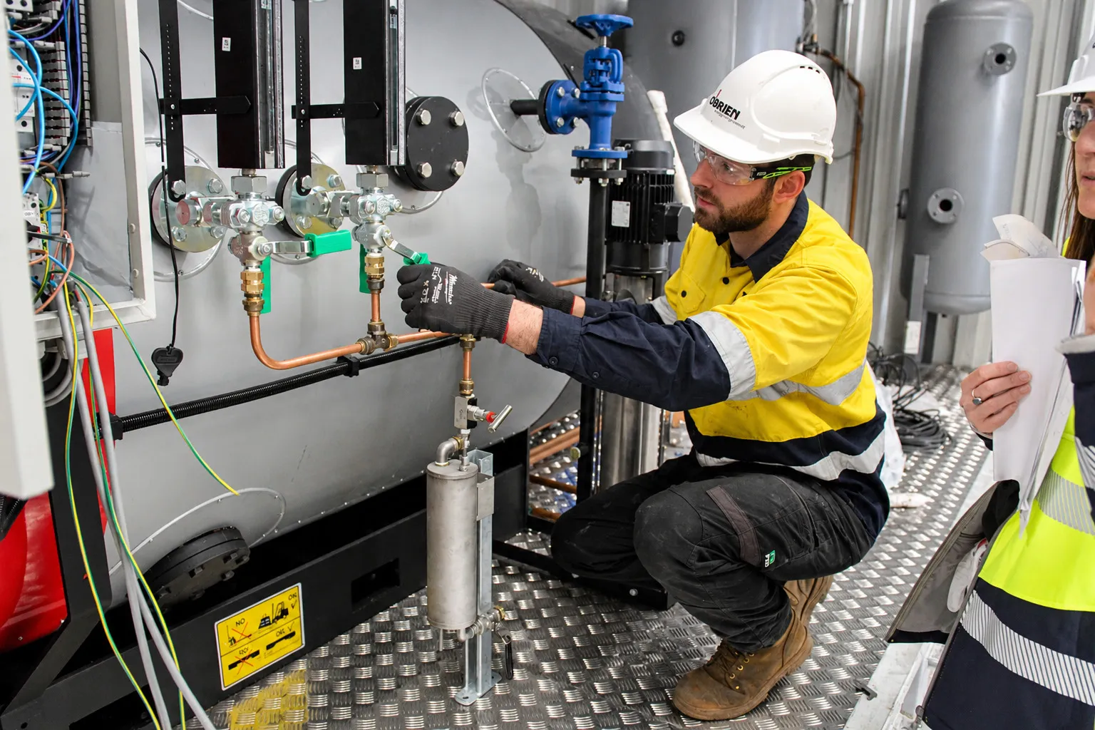 Boiler technician working a valve beside the water level gauges while a trainee takes notes
